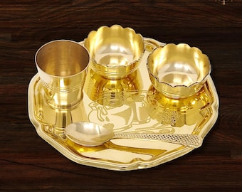 Op de afbeelding: Een gouden metalen puja thali set. De set bevat een dienblad, een kleine beker, twee geschulpte kommen en een lepel. De lade heeft geëtste ontwerpen en een geschulpte rand. De items zijn gerangschikt op een donker houten oppervlak.