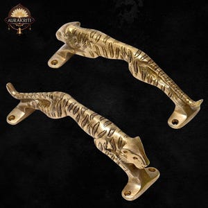 May include: Two brass tiger-shaped door handles. The handles are detailed with stripes and facial features, and are mounted on rectangular bases with screw holes. The handles are in a golden color and are set against a dark background. The brand name "AURAKRITI" is visible.