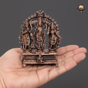 May include: Bronze-colored religious statue depicting three figures with ornate detailing. The central figure holds a bow and arrow, flanked by two others. The statue is held in a hand, showcasing its size and intricate design. The logo "AURAKRITI" is visible.