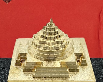 May include: A gold-colored, tiered pyramid sculpture with a square base. The sculpture has intricate details and a textured surface. The background is a solid red color. The logo "AURAKRITI" is visible in the top left corner.