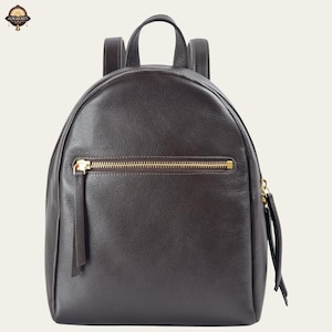 Könnte beinhalten: Ein dunkelbrauner Lederrucksack mit runder Form. Er hat einen goldfarbenen Reißverschluss und eine kleine Vordertasche. Der Rucksack hat zwei verstellbare Schultergurte und einen oberen Griff.