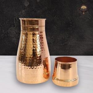 Könnte beinhalten: Ein kupferfarbenes Krug- und Becher-Set. Der Krug hat eine gehämmerten Textur und einen breiten Boden, der sich zu einem schmaleren Hals verjüngt. Der Becher ist ein kleinerer, passender Kupferbecher. Der Hintergrund ist eine dunkle, strukturierte Oberfläche.