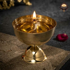 Könnte beinhalten: Eine brennende Öllampe aus Messing mit einer Flamme, die auf einer Steinoberfläche steht. Die Lampe hat eine runde Schalenform mit einem Sockel. Die Flamme ist hell, und die Lampe ist mit Öl gefüllt. Ein rotes Rosenblatt befindet sich im Hintergrund.