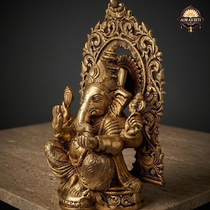 May include: A metallic gold-toned brass statue of the deity Ganesha, depicted in a seated position with four arms. The figure features an elephant head, ornate jewelry, and a textured dhoti, set against an intricate, arched, filigree-style backdrop.