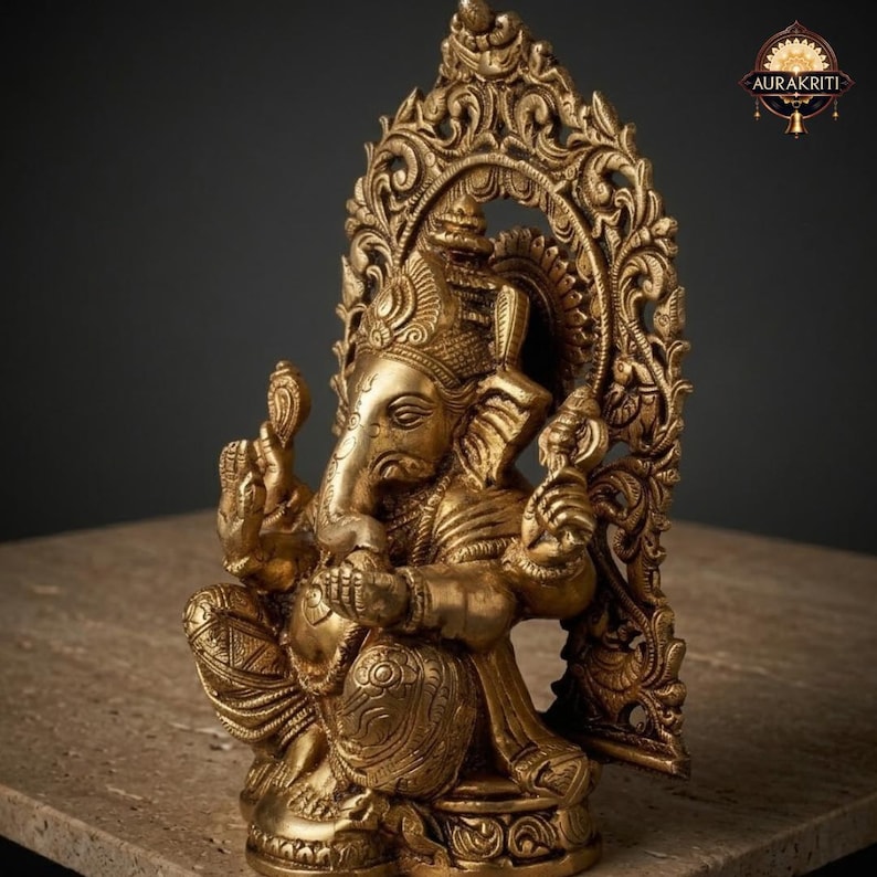 May include: A metallic gold-toned brass statue of the deity Ganesha, depicted in a seated position with four arms. The figure features an elephant head, ornate jewelry, and a textured dhoti, set against an intricate, arched, filigree-style backdrop.