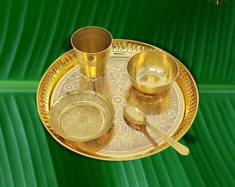 Op de afbeelding: Een goudkleurige puja thali set van messing met een beker, een kom, een klein bord, een lepel en een decoratief dienblad. Het dienblad en het bord hebben bloempatronen. De set staat op een groene achtergrond.