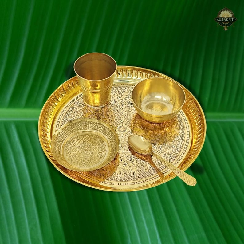 May include: A gold-colored brass puja thali set with a cup, bowl, small plate, spoon, and a decorative tray. The tray and plate feature floral patterns. The set is displayed on a green background.