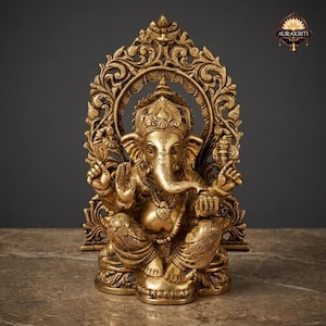 May include: A gold-toned brass statue of the deity Ganesha seated in a cross-legged position. The figure features an elephant head, multiple arms, and ornate jewelry. It is framed by an intricate, arched backdrop with floral carvings.