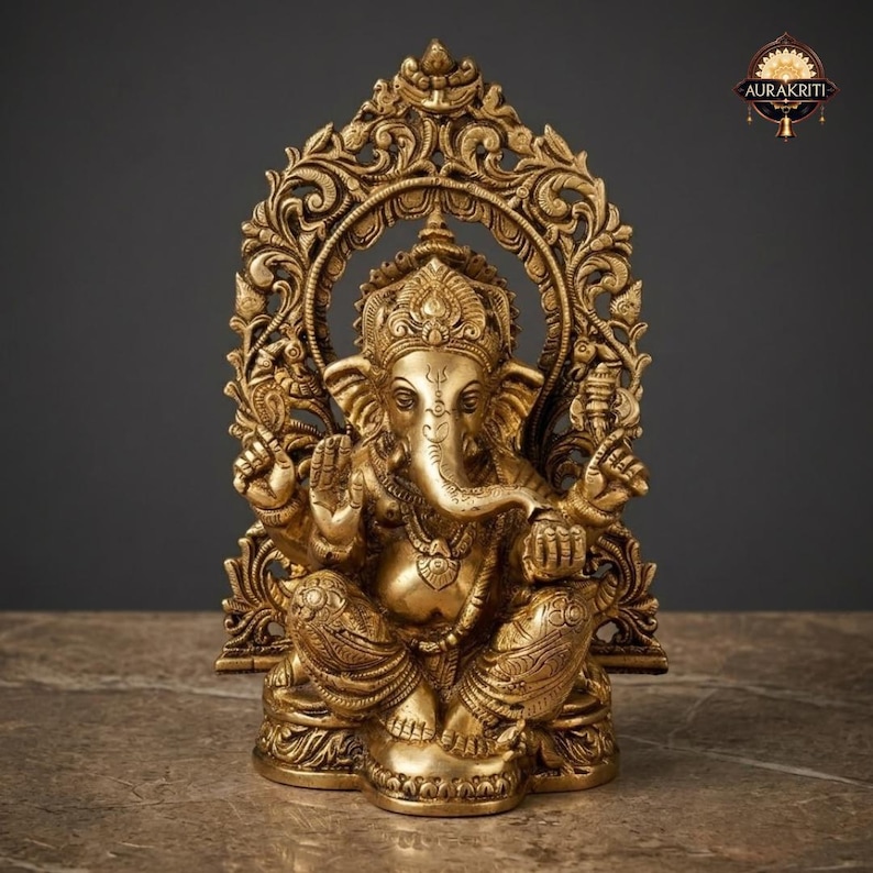 May include: A gold-toned brass statue of the deity Ganesha seated in a cross-legged position. The figure features an elephant head, multiple arms, and ornate jewelry. It is framed by an intricate, arched backdrop with floral carvings.