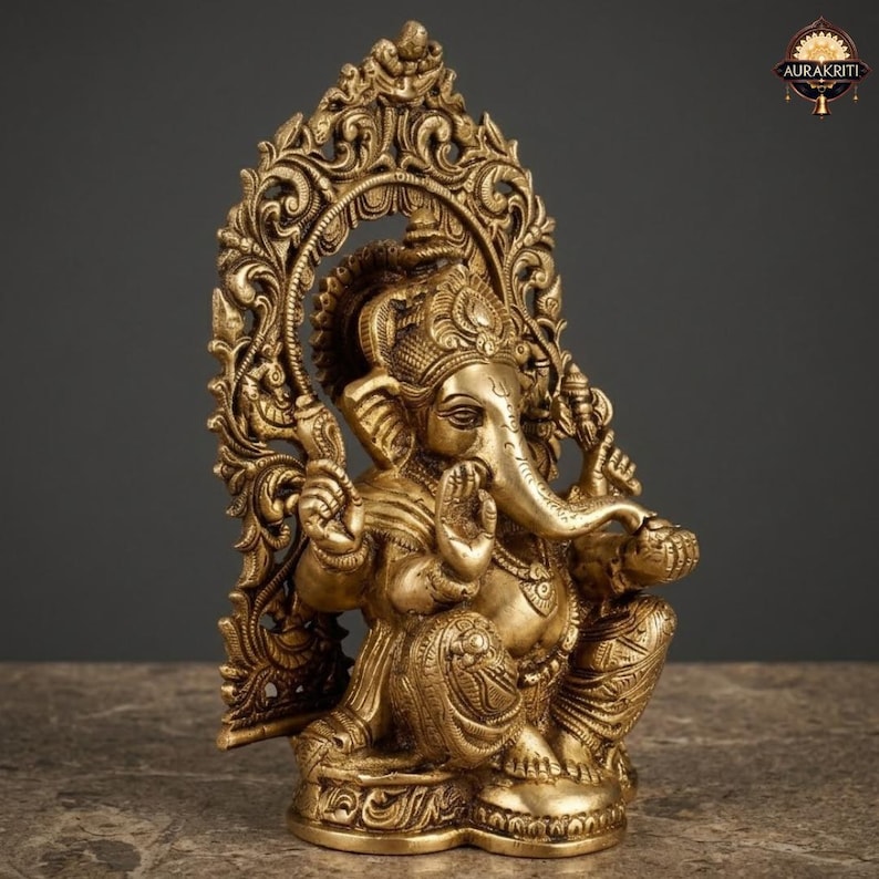 May include: A brass statue of the deity Ganesha depicted in a seated position with four arms, wearing an ornate crown and jewelry. The figure is framed by an intricate, arched backdrop featuring detailed floral and scrollwork carvings throughout.