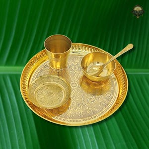 May include: A gold-colored brass puja thali set with a cup, small bowl, spoon, and small plate. The thali and its components have intricate floral patterns. The set is displayed on a green leaf background.