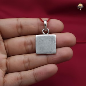 May include: A square silver pendant with a loop for a chain, suspended from a red cord. The pendant is held in a hand against a red background. The pendant has a simple, unadorned design.