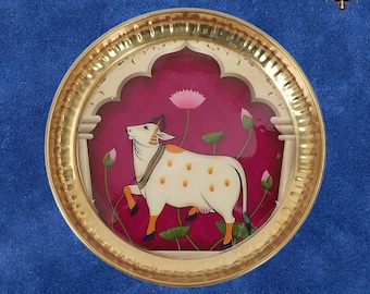 May include: A decorative round brass plate with a hammered texture. The plate features a painted scene of a white cow with orange spots, lotus flowers, and green leaves against a magenta background. The plate has a gold rim and a white arch design.