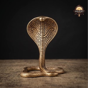 Könnte beinhalten: Eine Messing-Kobra-Statue mit detailliertem, strukturiertem Design. Die Schlange befindet sich in einer aufrechten Pose, mit aufgestelltem Kopf. Die Statue steht auf einer Holzoberfläche mit dunklem Hintergrund. Die Statue ist etwa 20 cm hoch.