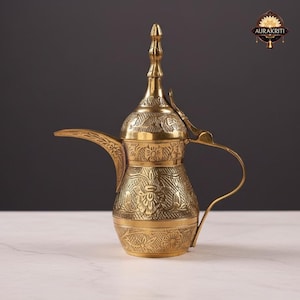 Könnte beinhalten: Eine goldfarbene, verzierte Kaffeekanne mit aufwendigen floralen und geometrischen Mustern. Die Kanne hat einen langen Ausguss, einen gebogenen Griff und einen dekorativen Deckel mit einer spitzen Spitze. Das Design deutet auf eine orientalische oder antike Ästhetik hin.