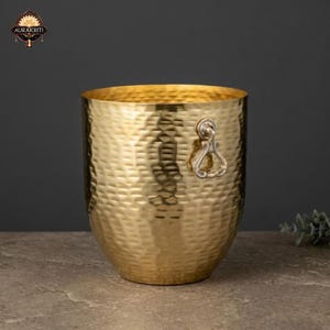 Puede incluir: Un cubo de metal de color dorado con una textura martillada y un cuerpo redondeado. El cubo cuenta con un asa de anillo metálico unida al lateral. El logotipo de Aurakriti es visible en la esquina superior izquierda de la imagen.
