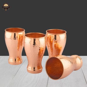 Puede incluir: Cuatro vasos de color cobre con una textura martillada. Tres vasos están de pie, mientras que uno está tumbado de lado. Los vasos tienen un cuerpo ancho y redondeado y una base acampanada. El interior de los vasos es de un color cobre más oscuro.