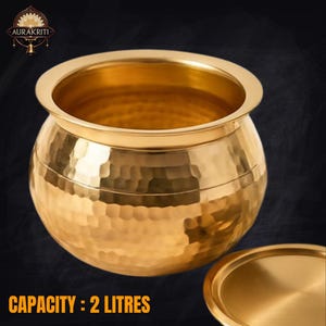 May include: A hammered brass pot with a rounded body and a wide rim. The pot has a capacity of 2 liters. The pot is gold in color and has a matching lid. The text "AURAKRITI" is in the upper left corner.