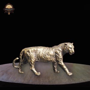 Op de afbeelding: Een bronzen tijgersculptuur in een wandelhouding. De sculptuur heeft een gedetailleerde vachttextuur en staat op een donker, rond oppervlak. Het logo "AURAKRITI" staat in de linkerbovenhoek.