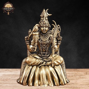 Può includere: Una statua in ottone dettagliata di una divinità seduta, adornata con gioielli intricati e un copricapo con una luna crescente. La figura tiene oggetti simbolici ed è seduta su una base a forma di loto. La statua è su una superficie di legno.