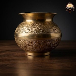 Puede incluir: Una olla de latón dorada y ornamentada, con boca ancha y cuerpo redondeado. La olla presenta intrincados diseños florales grabados en su superficie. El diseño incluye una banda horizontal en el centro. La olla está sobre una superficie de madera.