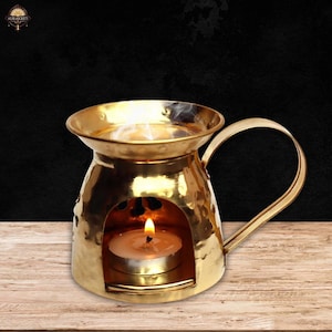 Könnte beinhalten: Ein goldfarbener Diffusor für ätherische Öle mit Griff und einer kleinen brennenden Kerze im Inneren. Der Diffusor hat eine gehämmerten Textur und eine runde Öffnung für die Kerze. Der Hintergrund ist schwarz und der Diffusor steht auf einer Holzoberfläche.