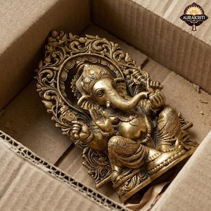 May include: A brass-colored metal statue of the deity Ganesha, depicted in a seated position with an elephant head and trunk, framed by an ornate, intricate archway featuring floral and scrollwork patterns, with the text AURAKRITI visible.
