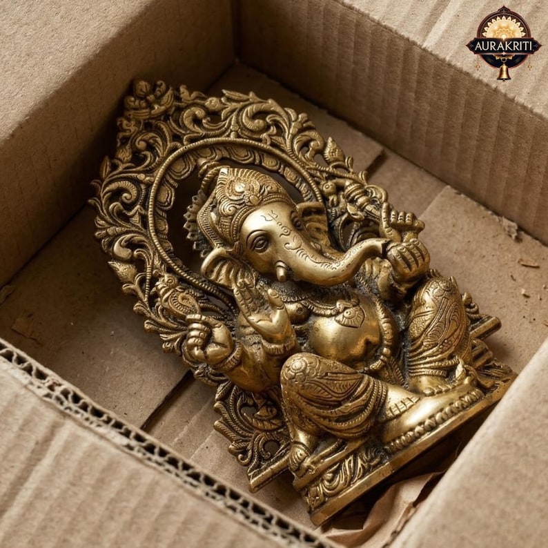 May include: A brass-colored metal statue of the deity Ganesha, depicted in a seated position with an elephant head and trunk, framed by an ornate, intricate archway featuring floral and scrollwork patterns, with the text AURAKRITI visible.