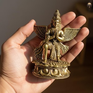 May include: A detailed brass statue of a winged deity, kneeling with hands in prayer. The figure has intricate detailing on the wings, clothing, and headdress. The statue is mounted on a decorative base and is held in a hand.