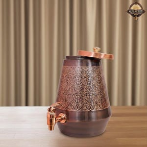 May include: Copper-colored water dispenser with an ornate floral pattern. The dispenser has a spigot and a lid with a crystal-like knob. The design is a mix of dark and light copper tones, creating a vintage aesthetic. The dispenser is on a wooden surface.