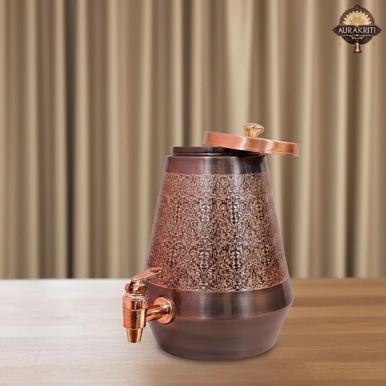 May include: Copper-colored water dispenser with an ornate floral pattern. The dispenser has a spigot and a lid with a crystal-like knob. The design is a mix of dark and light copper tones, creating a vintage aesthetic. The dispenser is on a wooden surface.