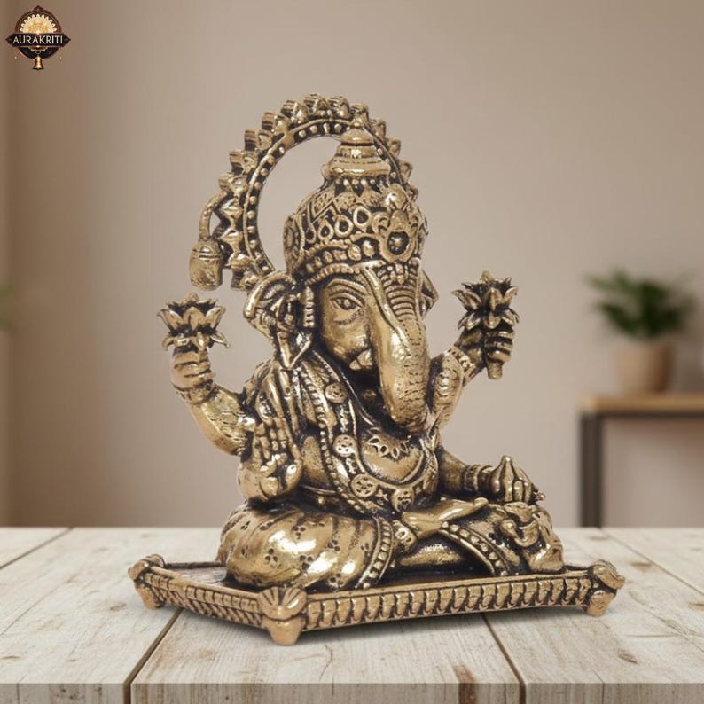 Może przedstawiać: Szczeg&oacute;łowa mosiężna statua hinduskiego b&oacute;stwa Ganesha, siedzącego w medytacyjnej pozie. Statua posiada skomplikowane rzeźbienia, trzyma kwiaty lotosu i jest zamontowana na dekoracyjnej podstawie. Widoczny jest tekst "AURAKRITI".