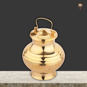 Puede incluir: Una olla de metal dorada, de forma redonda, con tapa y asa. La olla tiene estrías horizontales y se asienta sobre una pequeña base. El asa se arquea sobre la tapa. El fondo es un degradado de gris y negro.