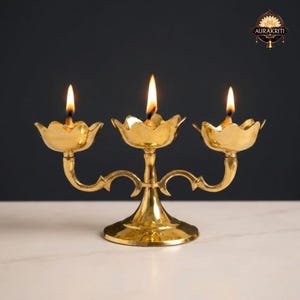 May include: A brass candle holder with three flower-shaped cups, each holding a lit candle. The holder has an ornate design with curved arms and a central stem. The flames are bright against a dark background.