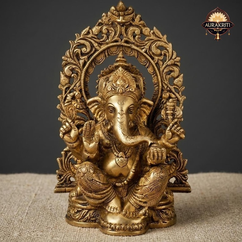 May include: A brass statue of the deity Ganesha, depicted in a seated position with four arms, an elephant head, and ornate jewelry. The figure is framed by an intricate, carved archway featuring floral motifs and the Aurakriti logo at the top.