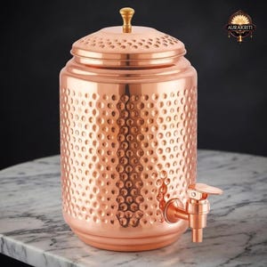 May include: A copper-colored water dispenser with a hammered texture. The dispenser has a lid with a gold-colored knob and a spigot. The product is cylindrical and sits on a marble surface. The brand name "AURAKRITI" is visible in the upper right corner.