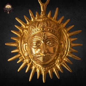 Könnte beinhalten: Ein goldfarbener Anhänger mit einem detaillierten Sonnengesicht mit Schnurrbart und Krone, umgeben von spitzen Strahlen. Der Anhänger hat oben eine Schlaufe zum Aufhängen. Das Logo "AURAKRITI" befindet sich in der oberen linken Ecke.