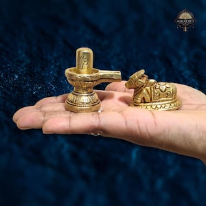 Op de afbeelding: Een paar kleine, goudkleurige religieuze beelden. Eén is een lingam, een cilindrische vorm, en de andere is een zittende stier. Beide zijn ingewikkeld gedetailleerd en rusten op een hand tegen een donkerblauwe achtergrond.