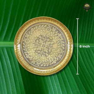 May include: A gold-colored, round decorative plate with an intricate floral pattern. The plate is 8 inches in diameter and has a detailed border. The background is a vibrant green, creating a striking contrast. The plate is likely made of metal.