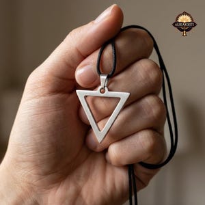 Könnte beinhalten: Halskette mit silberfarbenem Dreiecksanhänger und schwarzem Band. Das offene Dreieck-Design wird in einer Hand gehalten. Die Halskette hat eine schlichte, moderne Ästhetik. Das Markenlogo ist oben rechts sichtbar.