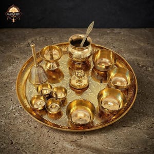 May include: A polished brass puja thali set featuring a large circular tray with an intricate floral engraving, a bell, a small pot with a spoon, a diya lamp, a central incense holder, and several small bowls for offerings, all in a metallic gold finish.