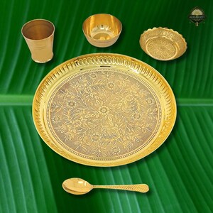 May include: A collection of gold-colored serving pieces, including a large plate with floral detailing, a small bowl, a cup, a decorative dish, and a spoon. The items are arranged on a green leaf background, showcasing the intricate designs and the warm tones of the metal.