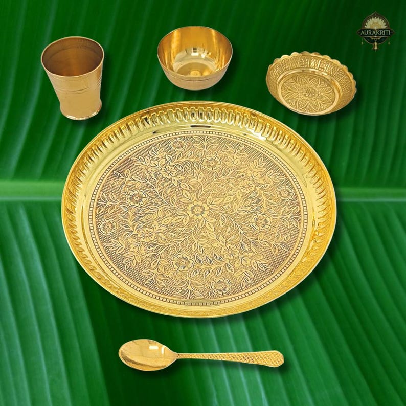 May include: A collection of gold-colored serving pieces, including a large plate with floral detailing, a small bowl, a cup, a decorative dish, and a spoon. The items are arranged on a green leaf background, showcasing the intricate designs and the warm tones of the metal.