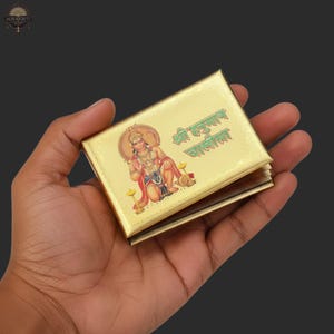 Könnte beinhalten: Ein goldfarbenes, rechteckiges Buch mit einer Illustration einer Gottheit und grünem Text. Das Buch wird in einer Hand vor einem dunklen Hintergrund gehalten. Die Illustration zeigt eine Gottheit in roter und goldener Kleidung.