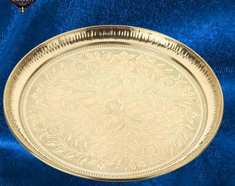 May include: A gold-colored, oval-shaped serving tray with intricate floral patterns. The tray has a detailed border and a polished finish. The background is a deep blue fabric. The tray is likely used for serving food or displaying decorative items.