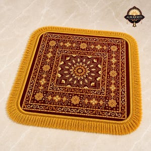 Puede incluir: Una alfombrilla cuadrada de color granate con un patrón central de mandala floral dorado, rodeada de intrincados bordes geométricos y un grueso fleco dorado. El diseño incluye el logotipo de Aurakriti en la esquina superior derecha en oro y negro.