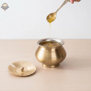 Może przedstawiać: Mosiężny garnek ze srebrną obręczą i pasującą pokrywką. Łyżka wlewa złoty płyn do garnka. Garnek stoi na jasnej drewnianej powierzchni.