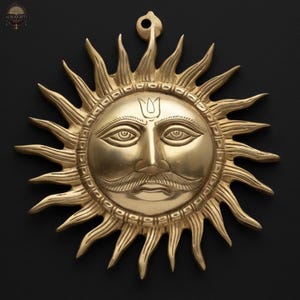 May include: A golden sun-shaped wall hanging with a detailed face and radiating rays. The sun has a mustache and a decorative mark on its forehead. The piece is made of metal and has a loop for hanging. The background is black.