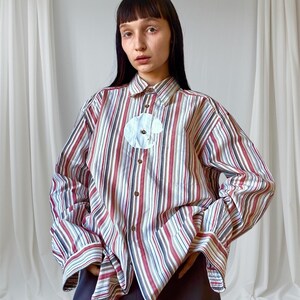 May include: A striped button-down shirt with vertical stripes in red, white, and blue. The shirt features a white floral applique and long sleeves. The model is wearing a dark blue skirt.