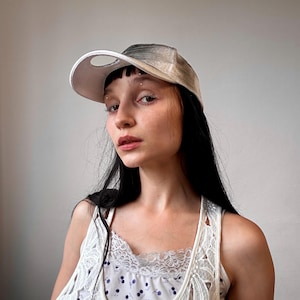 Puede incluir: Gorra de béisbol con visera blanca y corona gris y beige. La gorra tiene una abertura circular en la parte superior. La persona lleva una camiseta sin mangas blanca con un estampado floral.